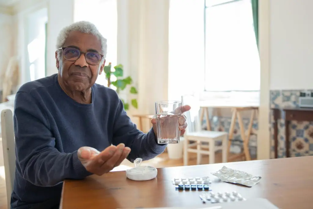 An older gentleman seated at a table with medication and water, considering the critical benefits of Vitamin D for well-being.