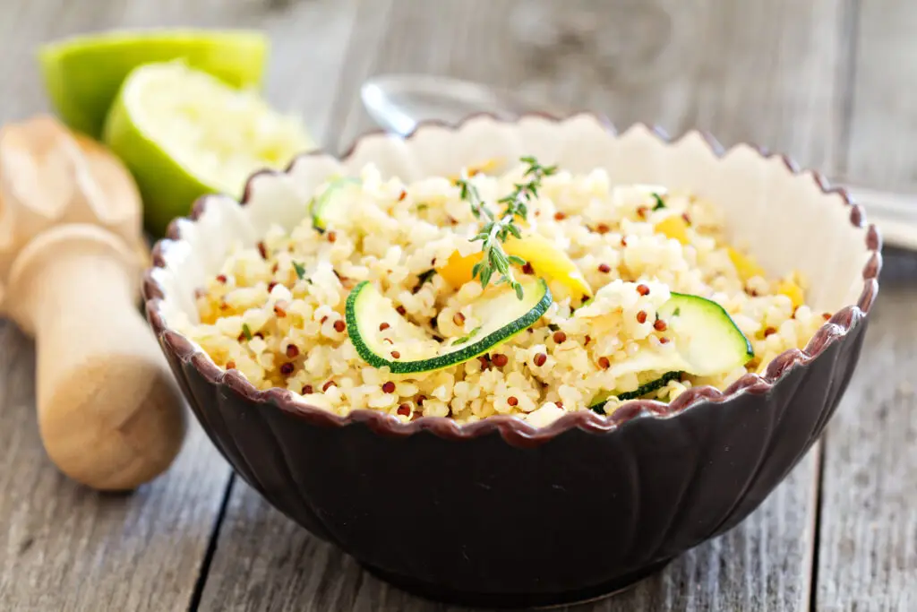 A bowl of quinoa salad with sliced courgettes and herbs, served on a rustic wooden table with a lime wedge in the background - a nutrient-dense dish supporting hormone balance and heart health.